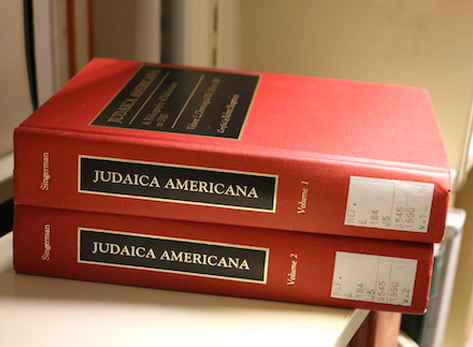 A photo of volumes 1 and 2 of Robert Singerman's JUDAICA AMERICANA stacked on a library shelf. The books have bright red ocvers and the title written in gold inside a black box.