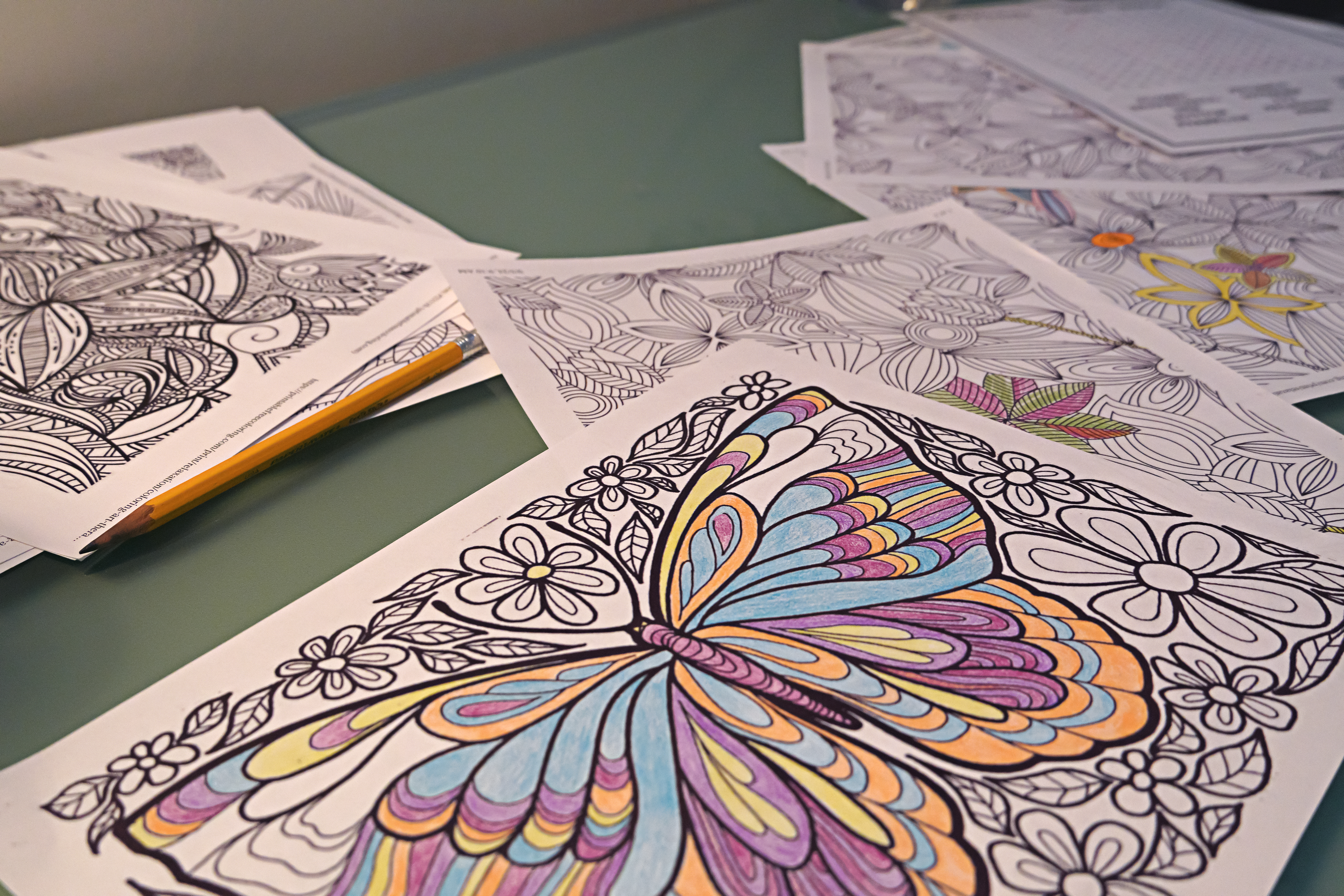 A variety of coloring sheets lay on a table. The top sheet depicts a butterfly and is partially colored in with crayon or marker.