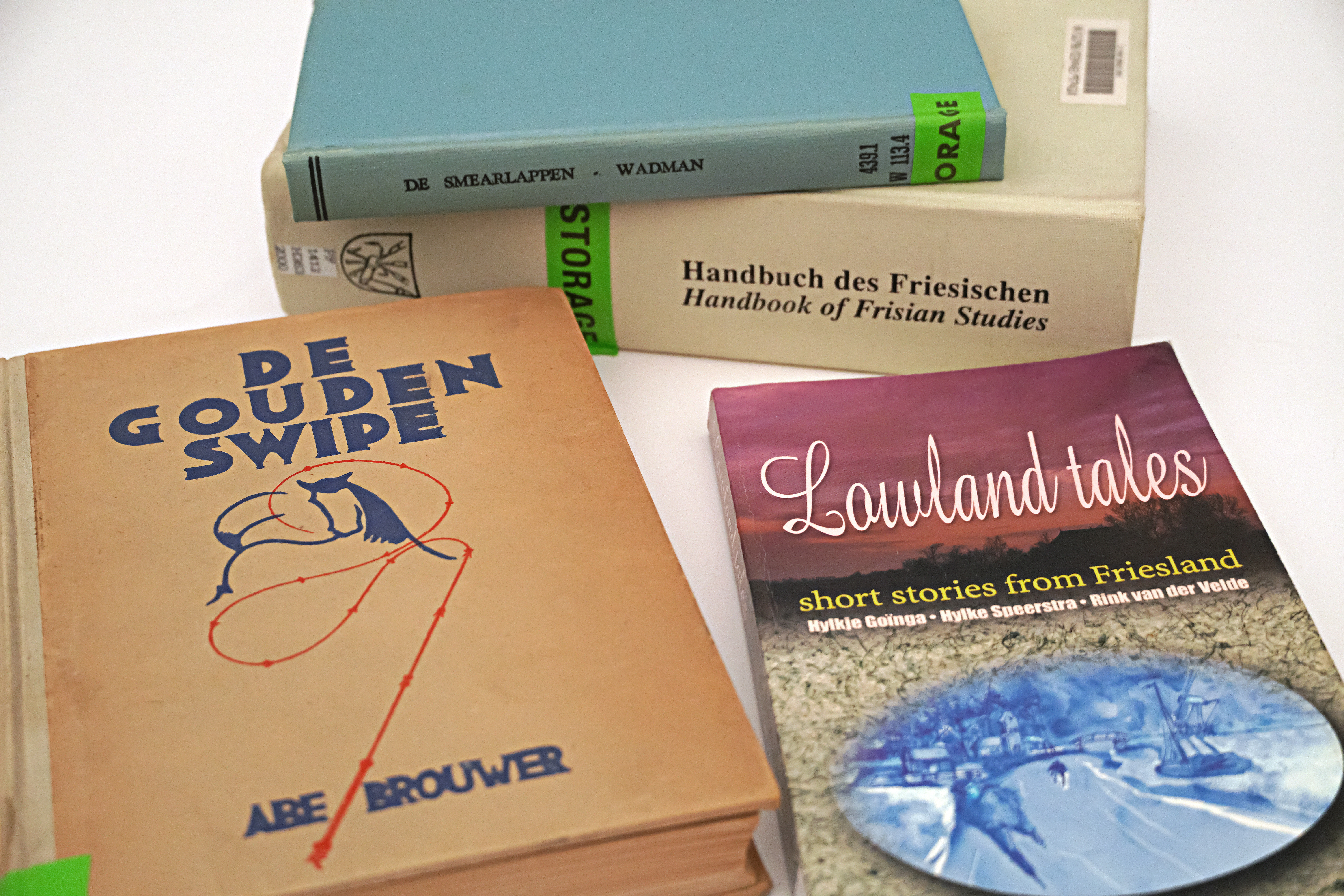 Four books arranged on white surface. The titles are mixtures of German, English, and both. De Gouden Swipe has a drawing of a horse and rider with a red whip. Lowland Tales features a landscape image.