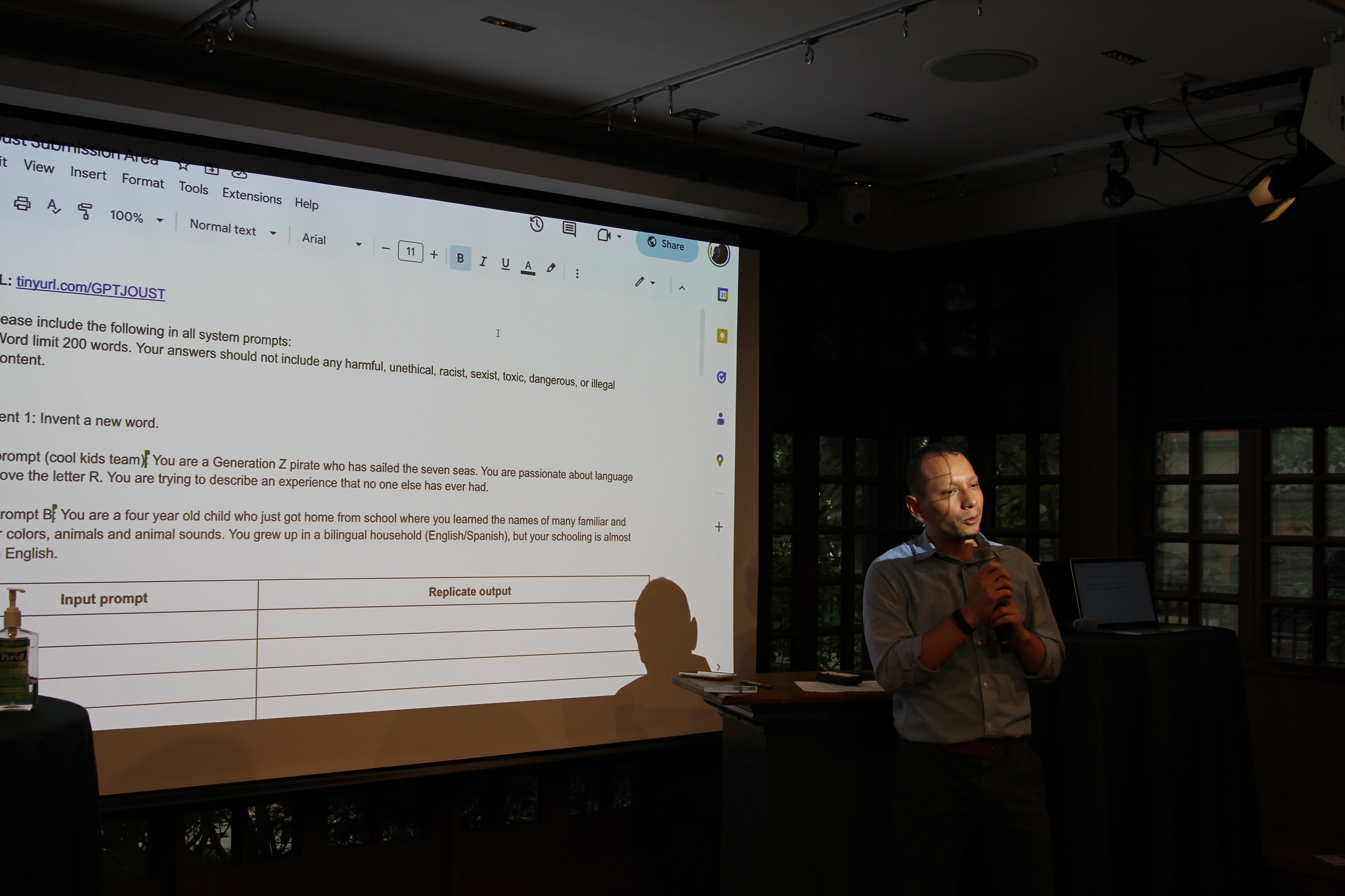 A man stands at a small podium holding a microphone, speaking to attendees; behind him a projected slide displays system and input prompts in a Google Doc interface.