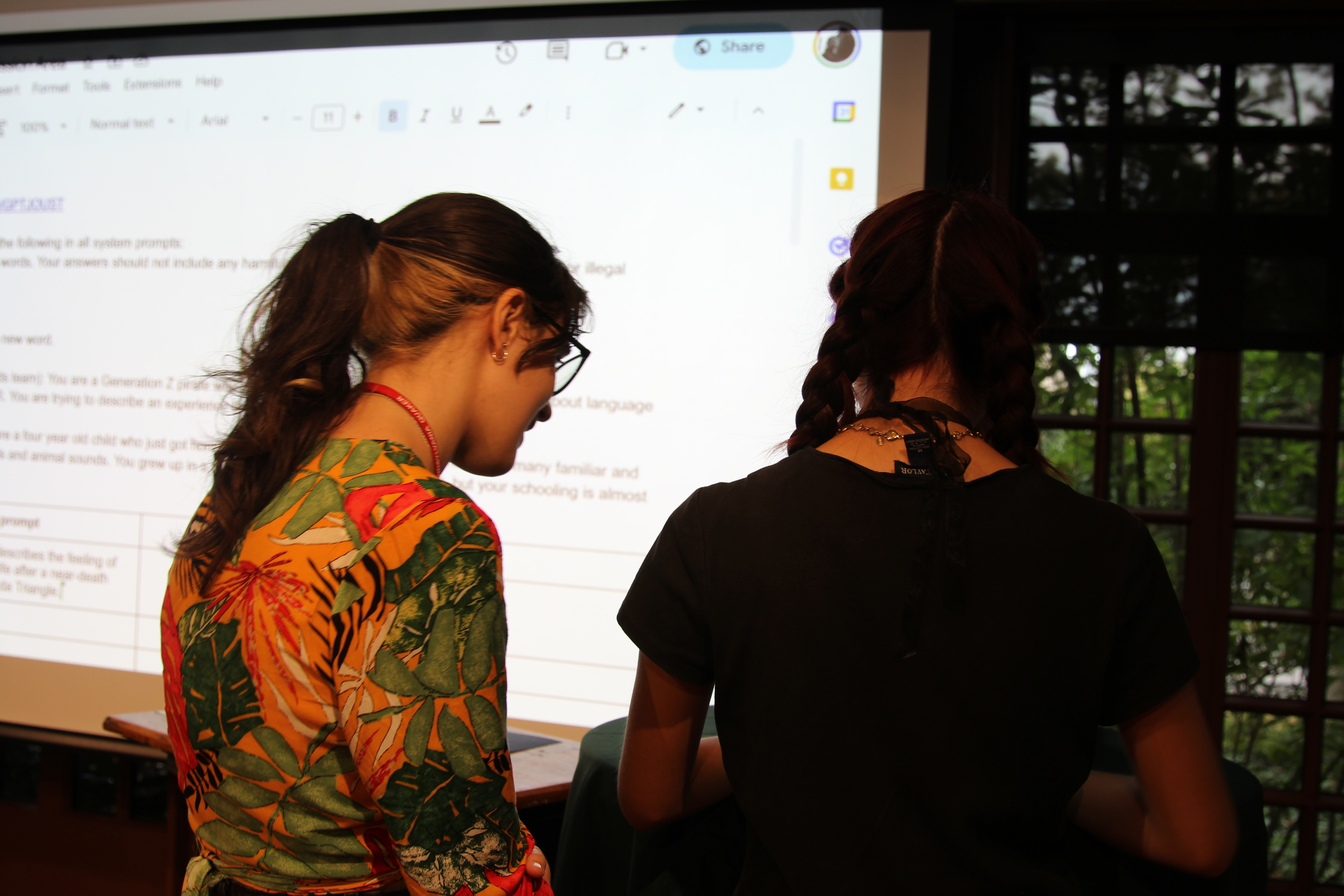 Two participants stand at the front of the room entering their custom prompts into the live Google Doc interface projected on the screen, collaborating quietly before the next round begins.