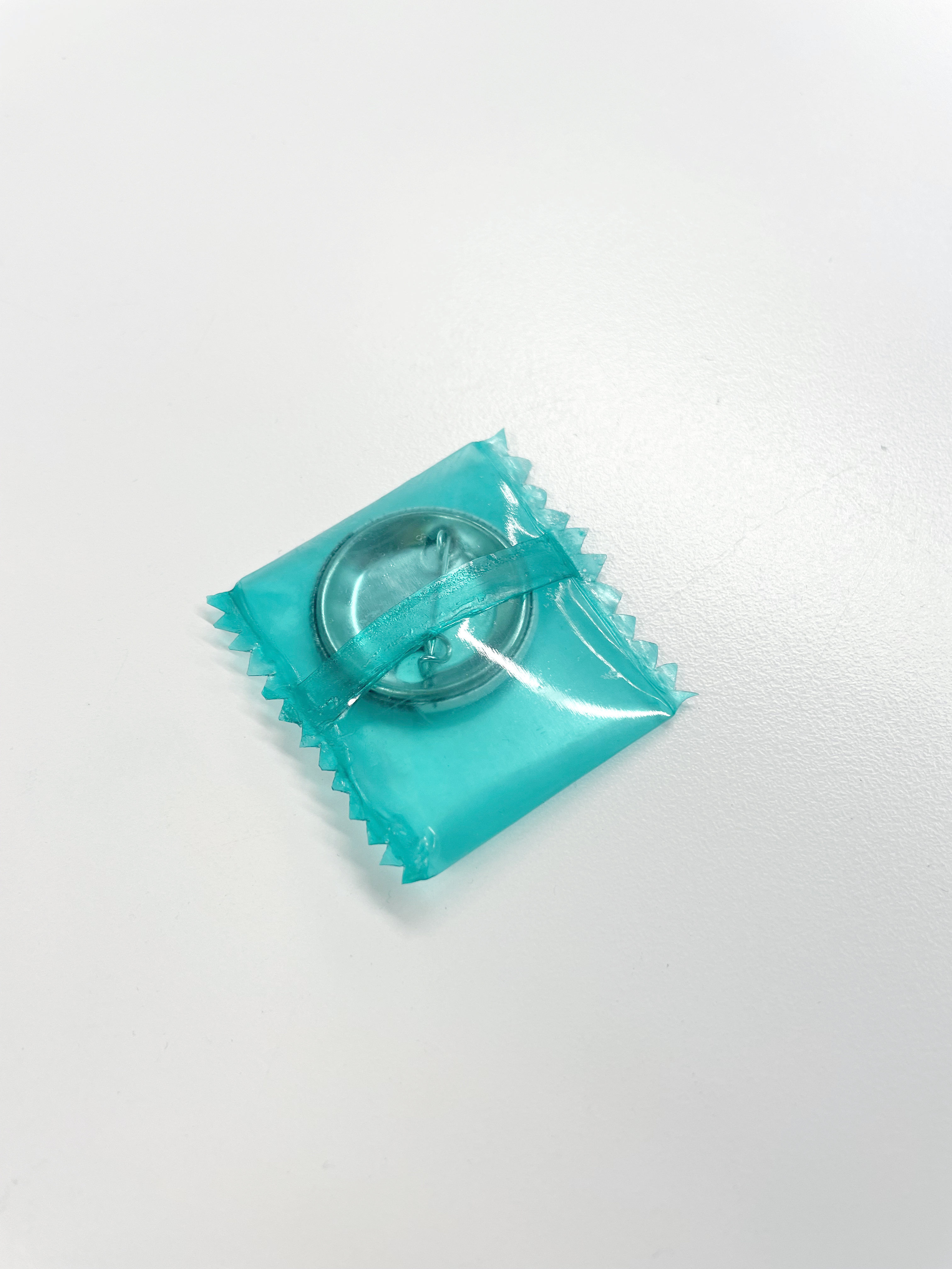  A clear, teal-tinted plastic wrapper with zigzag edges encloses a circular, metallic object. A matching teal ribbon is visible around the object.