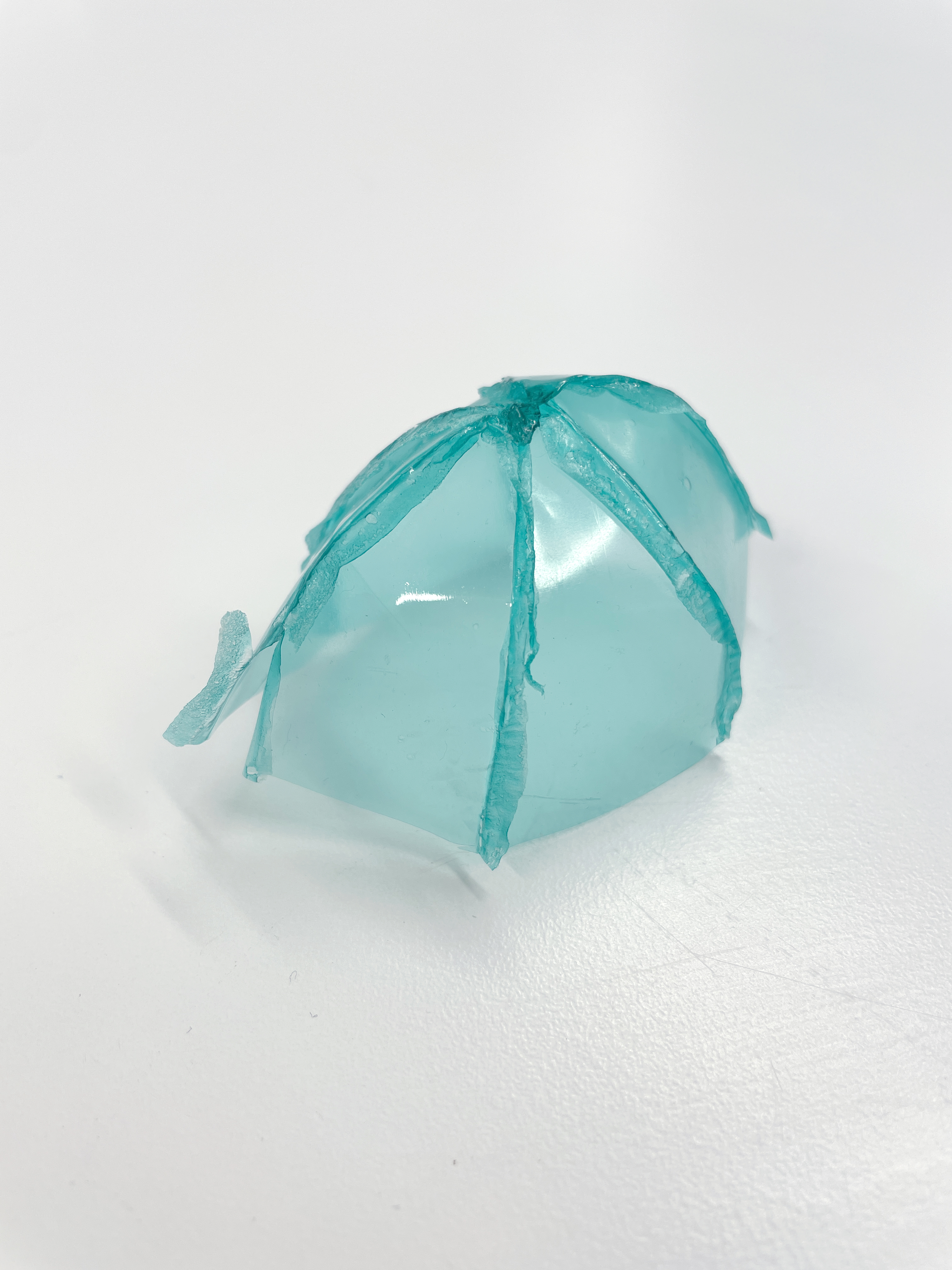  A transparent, teal, dome-shaped object with visible seams and a slightly irregular base, sitting on a white surface.
