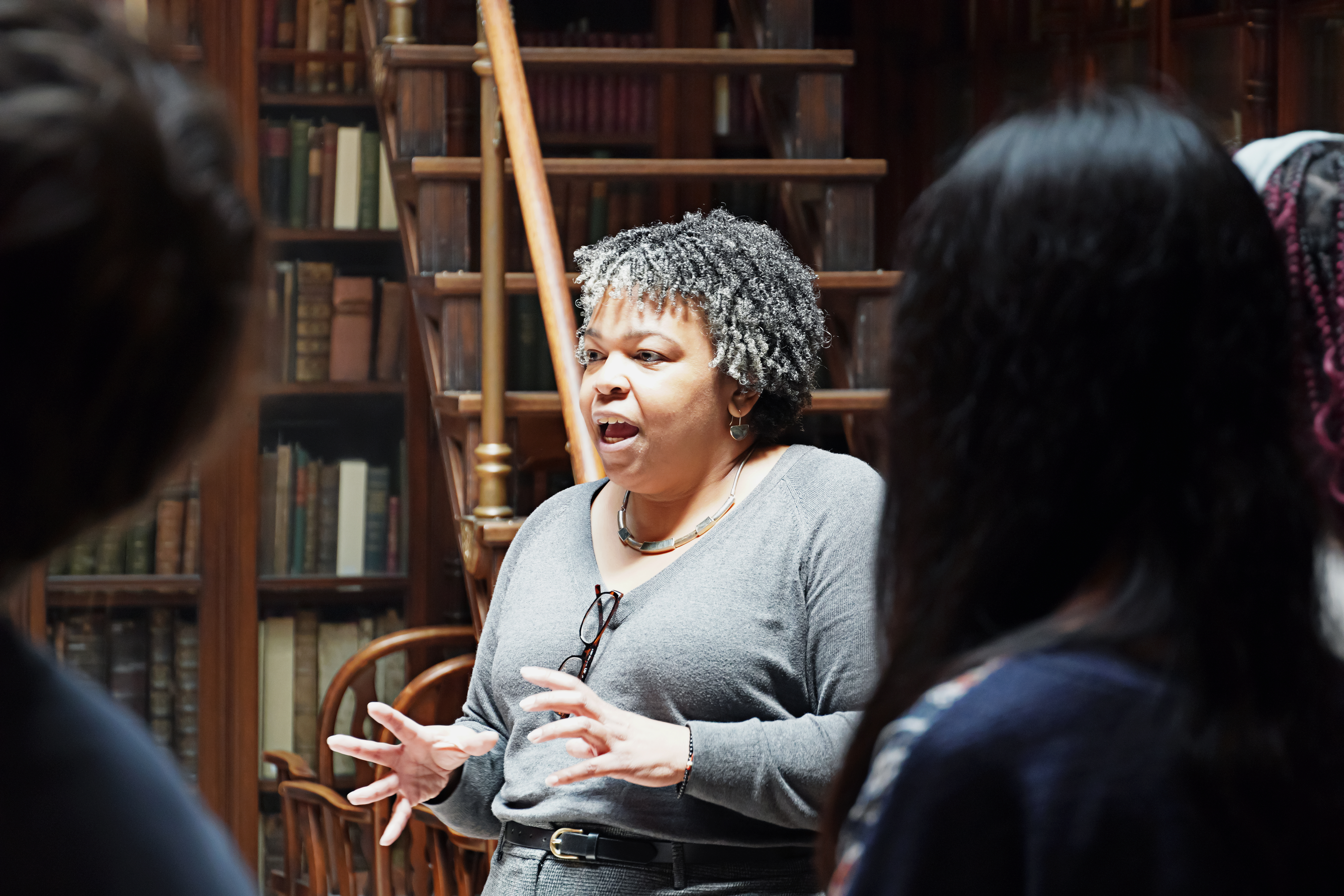 A woman in a library is speaking animatedly while gesturing with her hands. She is surrounded by books and a wooden staircase.