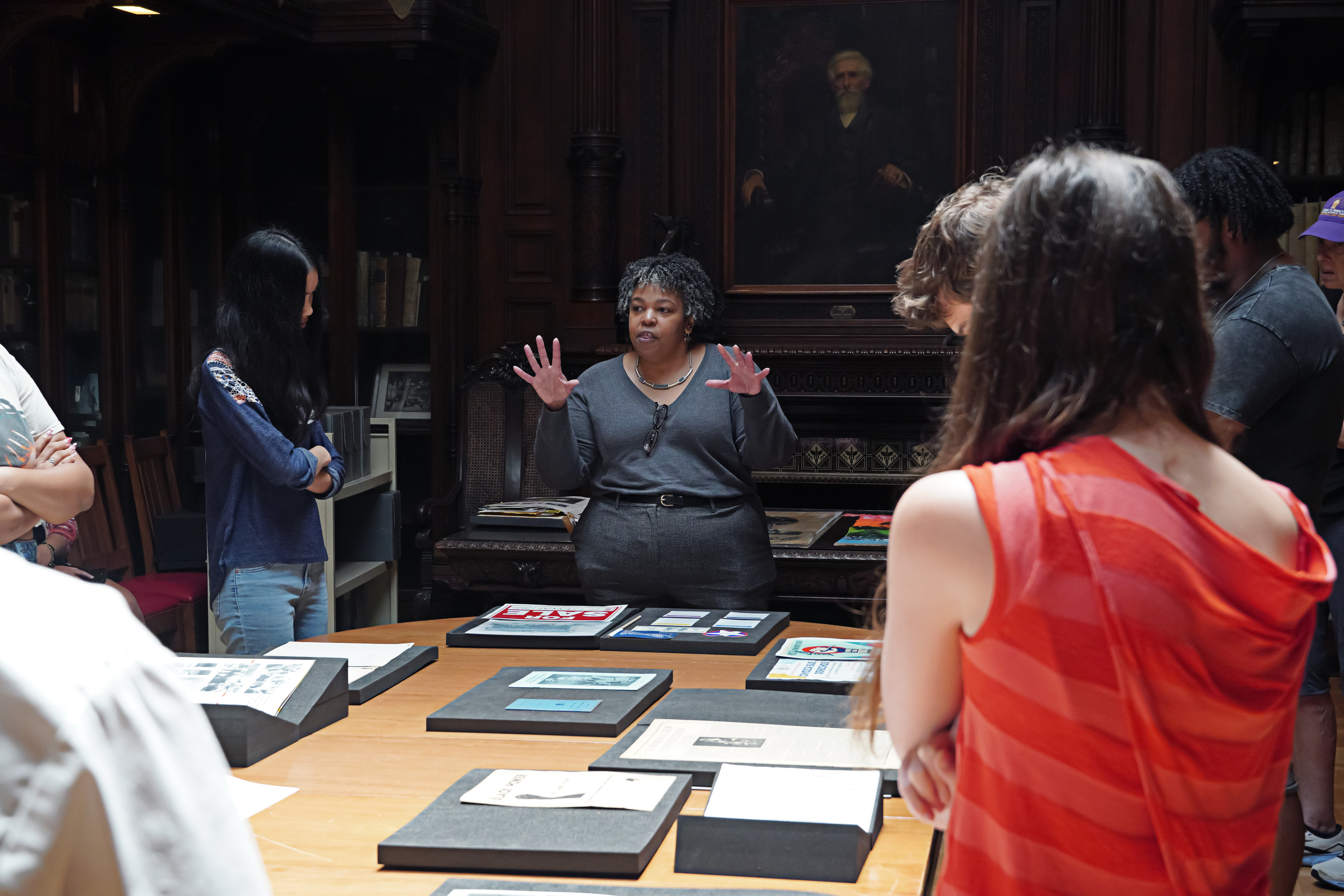  A librarian stands at the head of a table with documents on it, speaking to a group of students. A portrait hangs on the wall behind her.