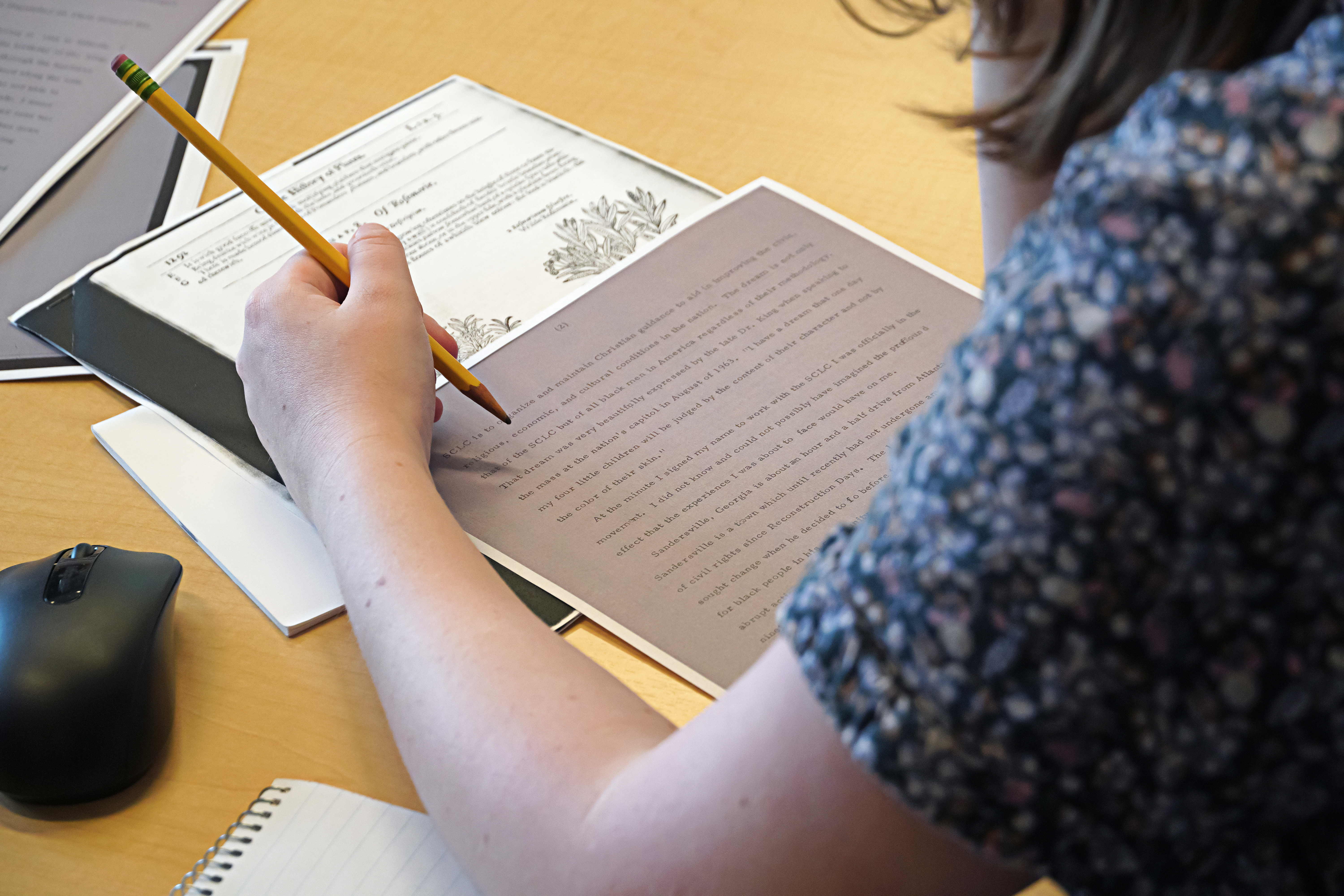  A student takes notes with a pencil on a document with text on a table. A computer mouse and a notebook are nearby.