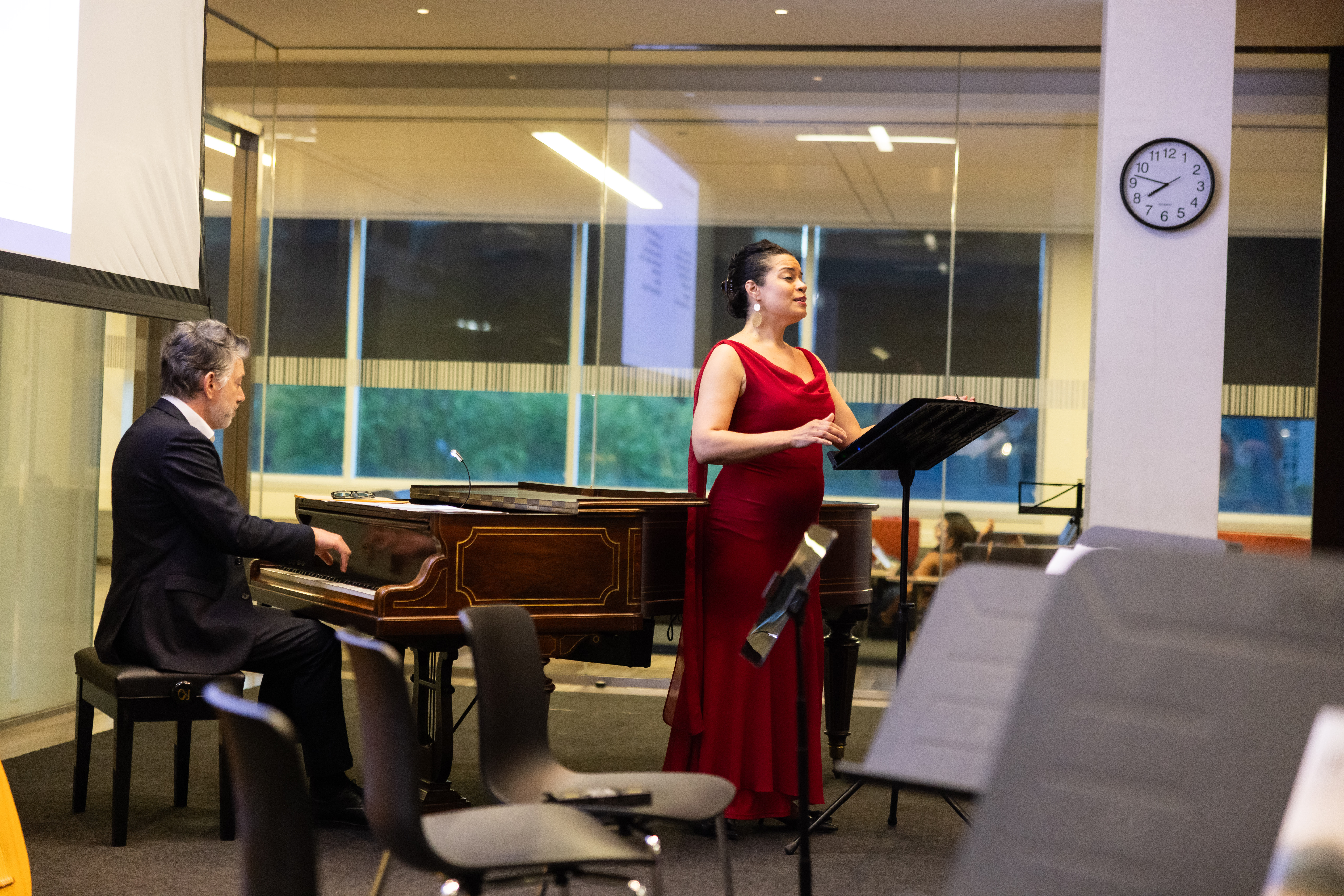 A person plays the piano while another person sings while standing in front of a music stand
