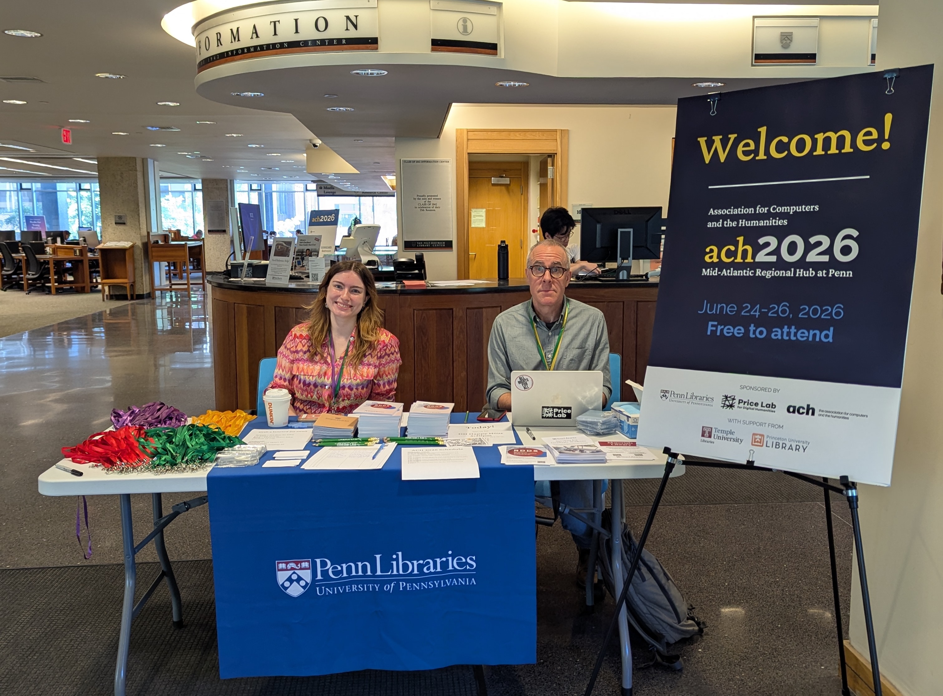 Adrianna Bugliarello-Wondrich and Stewart Varner sit behind a table with a Penn Libraries tablecloth in the first-floor entryway to Van Pelt Library. A sign on an easel to the right of the table bids a welcome to the ACH2026 Mid-Atlantic Regional Hub.