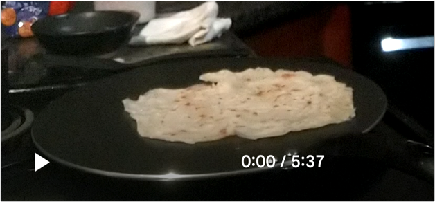 Flatbread cooking on a large round griddle on a stovetop, shown in a video player screenshot.