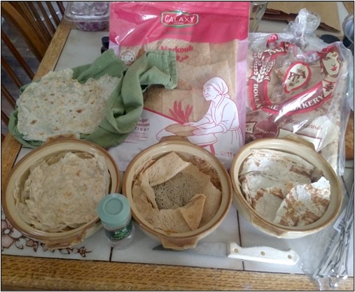 Preparation ingredients for making flatbread arranged on a table. Three bowls contain dough, torn pieces of dough, and flour-coated dough, with a small spice container in front. Several round flatbreads rest on a green cloth beside packages of flour and baking ingredients in the background. A rolling pin and utensils are visible at the edge of the table.