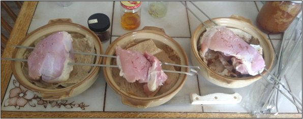 Three clay pots on a kitchen table, each holding ingredients topped with raw chicken pieces on metal skewers, with seasonings and cooking utensils nearby.