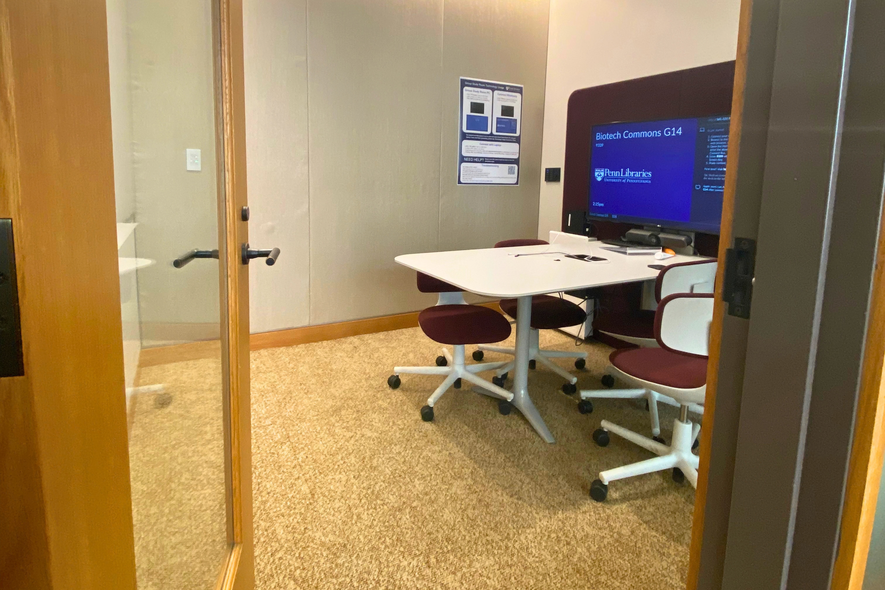 Small library group study room viewed through an open doorway, featuring a white conference table surrounded by rolling chairs with burgundy seats. A large wall-mounted display screen shows room information for “Biotech Commons G14” and Penn Libraries. The room has carpeted flooring, acoustic wall panels, and a glass door.