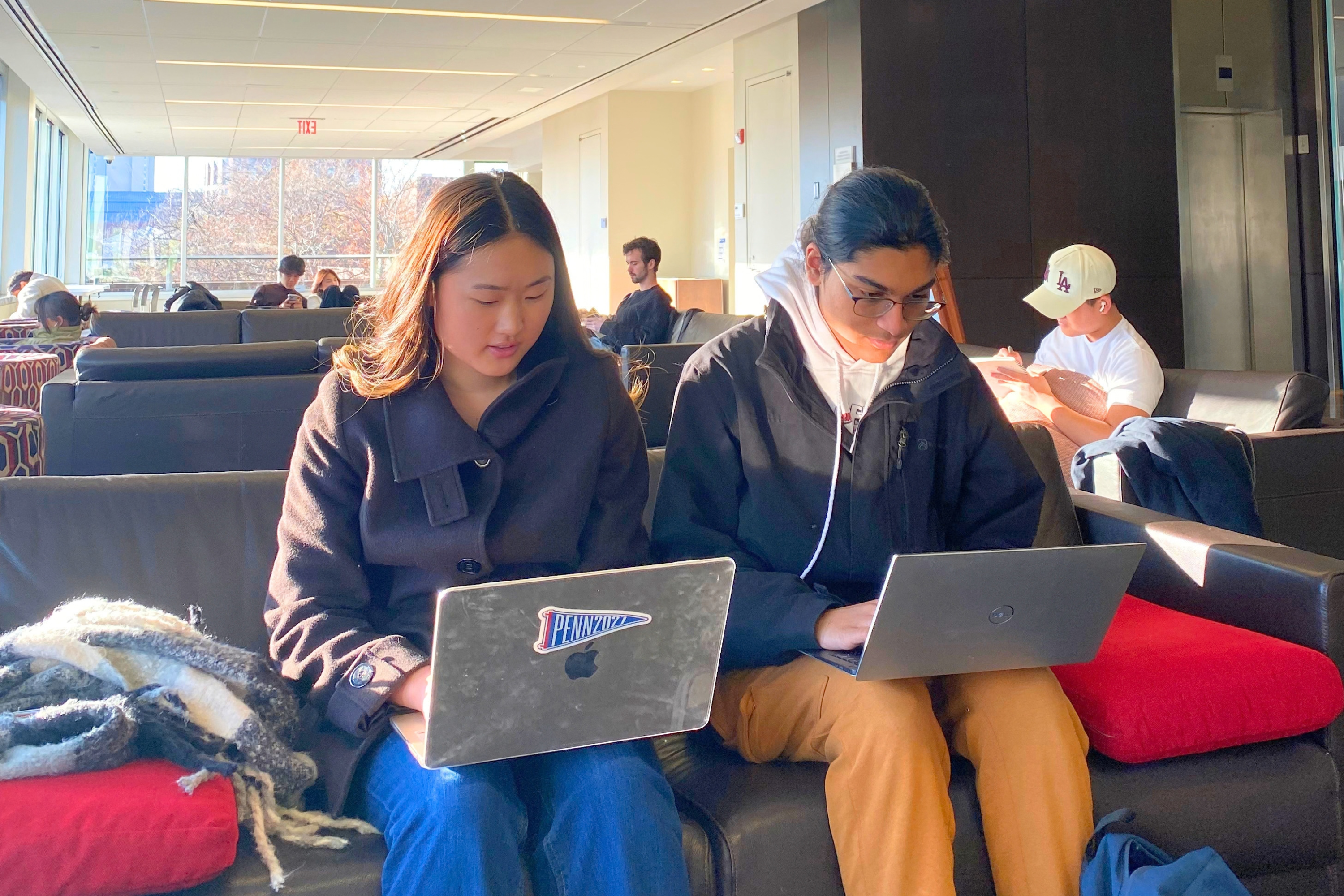 Students work on laptops while seated on cushioned sofas in a bright library lounge. Large windows fill the space with natural sunlight, and additional students study or use devices in the background.